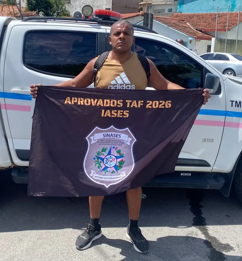 Edinardo segurando a bandeira Aprovados TAF 2026 IASES em frente a uma viatura.