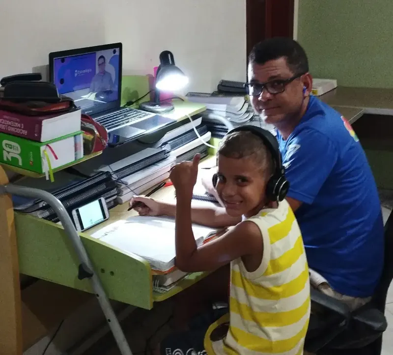 Hélio estudando em casa ao lado do filho, com livros, apostilas e uma videoaula aberta no notebook.